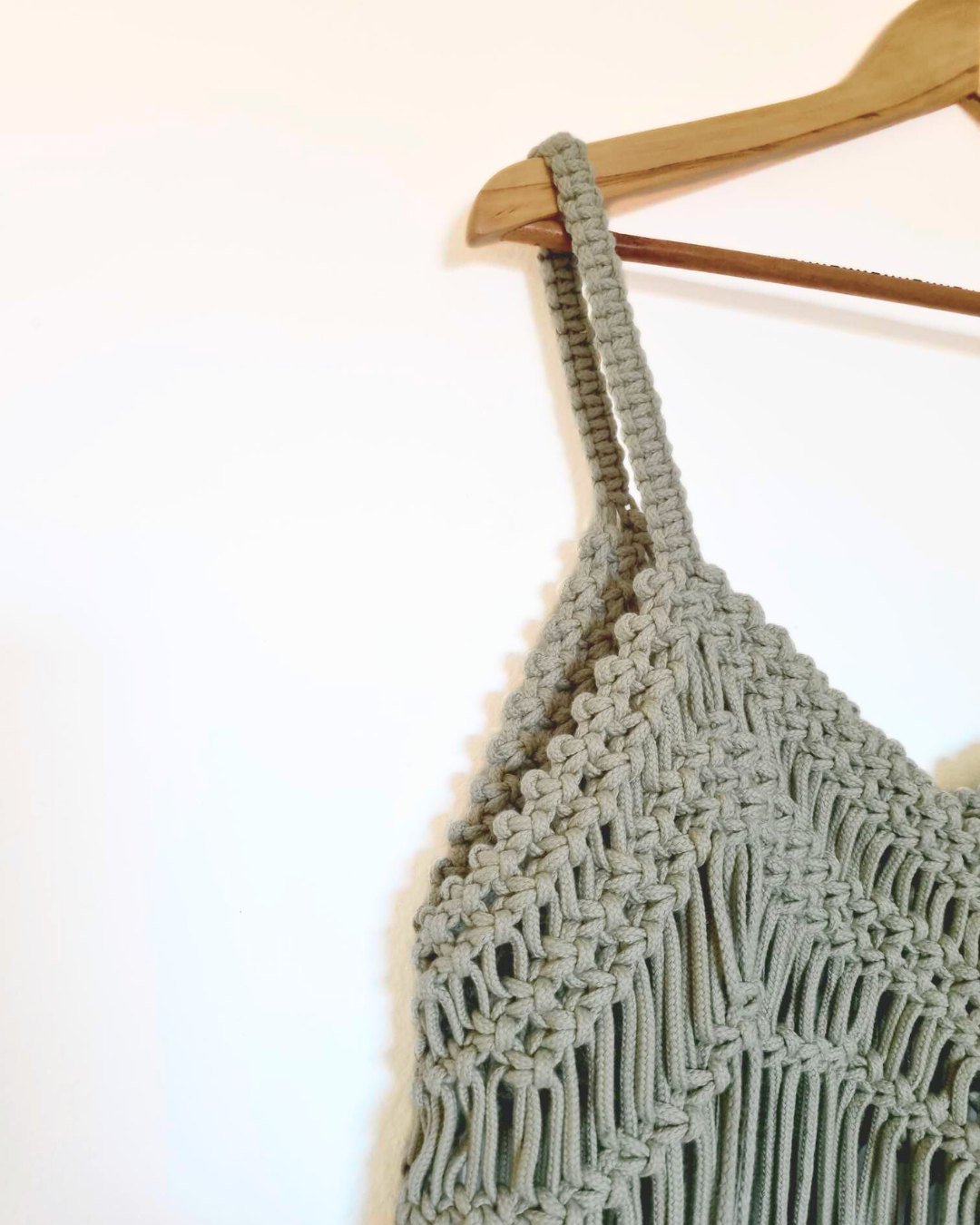 Detalhe de top em macramé verde-sage suspenso em cabide de madeira — peça artesanal feita à mão | Jungle Macramé